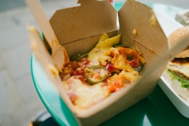 A close-up view of a takeaway box filled with nachos, topped with melted cheese, jalapeños, and diced tomatoes. Next to it, part of a hamburger with lettuce and sauce is visible, set on a plastic tray. The scene is situated on a green table, likely outside.