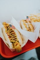 Two hot dogs are placed on a red tray, each topped with mustard, relish or onions, and garnished with small American flags. The hot dogs are served in paper liners. A portion of fries is visible in the background.