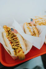 Two hot dogs are placed on a red tray, each topped with mustard, relish or onions, and garnished with small American flags. The hot dogs are served in paper liners. A portion of fries is visible in the background.