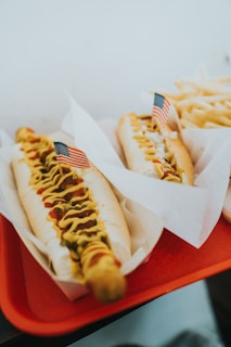 Two hot dogs are placed on a red tray, each topped with mustard, relish or onions, and garnished with small American flags. The hot dogs are served in paper liners. A portion of fries is visible in the background.
