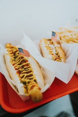 Two hot dogs are placed on a red tray, each topped with mustard, relish or onions, and garnished with small American flags. The hot dogs are served in paper liners. A portion of fries is visible in the background.