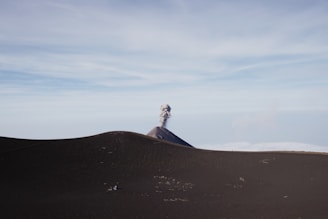 A distant volcano with a plume of smoke rising from its peak is set against a vast landscape with rolling hills or dunes in the foreground. The sky is clear and expansive, giving a sense of openness and isolation.