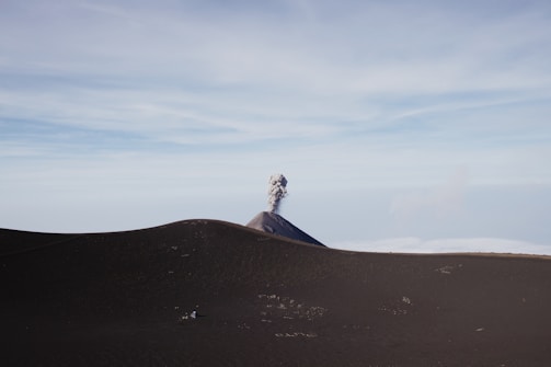 A distant volcano with a plume of smoke rising from its peak is set against a vast landscape with rolling hills or dunes in the foreground. The sky is clear and expansive, giving a sense of openness and isolation.