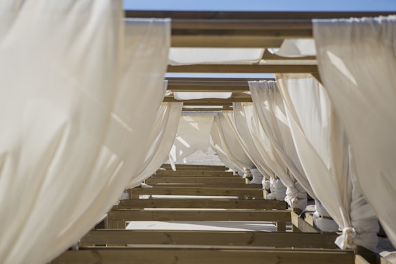 Rows of white fabric curtains hang from a structure of wooden beams, creating a rhythmic pattern of light and shadow. The fabric appears slightly translucent with a gentle breeze causing slight movement.