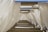 Rows of white fabric curtains hang from a structure of wooden beams, creating a rhythmic pattern of light and shadow. The fabric appears slightly translucent with a gentle breeze causing slight movement.