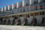 A multi-level residential building features several staircases leading up to each unit. The structure has a series of windows and railings. Decorative colorful streamers are strung across the building, adding a festive touch.