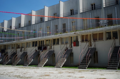 A multi-level residential building features several staircases leading up to each unit. The structure has a series of windows and railings. Decorative colorful streamers are strung across the building, adding a festive touch.
