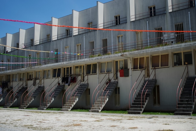 A multi-level residential building features several staircases leading up to each unit. The structure has a series of windows and railings. Decorative colorful streamers are strung across the building, adding a festive touch.