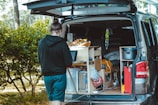 A person stands next to an open van with a customized interior featuring a small kitchen setup, including storage compartments and various supplies. The outdoor setting includes trees and greenery, suggesting a camping or travel scenario.