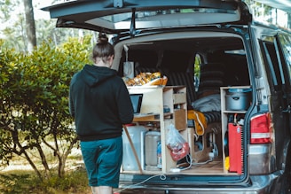 A person stands next to an open van with a customized interior featuring a small kitchen setup, including storage compartments and various supplies. The outdoor setting includes trees and greenery, suggesting a camping or travel scenario.