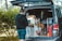 A person stands next to an open van with a customized interior featuring a small kitchen setup, including storage compartments and various supplies. The outdoor setting includes trees and greenery, suggesting a camping or travel scenario.