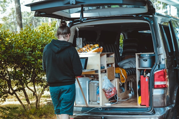 A person stands next to an open van with a customized interior featuring a small kitchen setup, including storage compartments and various supplies. The outdoor setting includes trees and greenery, suggesting a camping or travel scenario.