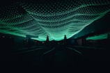 Green illuminated netting or mesh covers a dark walkway, casting an ambient glow in the surrounding area. The netting features a hexagonal pattern and appears to be suspended overhead, creating a dynamic and surreal visual effect.