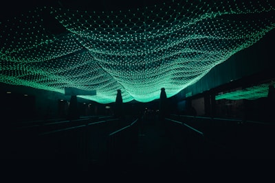 Green illuminated netting or mesh covers a dark walkway, casting an ambient glow in the surrounding area. The netting features a hexagonal pattern and appears to be suspended overhead, creating a dynamic and surreal visual effect.