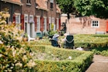 Two elderly people sit in a neatly manicured garden with chairs, enjoying a sunny day. The garden is surrounded by quaint brick houses with red shutters and lush greenery. Potted plants and flowers add color, while the sunlight casts gentle shadows.