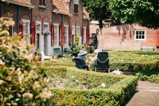 Two elderly people sit in a neatly manicured garden with chairs, enjoying a sunny day. The garden is surrounded by quaint brick houses with red shutters and lush greenery. Potted plants and flowers add color, while the sunlight casts gentle shadows.