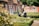 Two elderly people sit in a neatly manicured garden with chairs, enjoying a sunny day. The garden is surrounded by quaint brick houses with red shutters and lush greenery. Potted plants and flowers add color, while the sunlight casts gentle shadows.