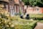 Two elderly people sit in a neatly manicured garden with chairs, enjoying a sunny day. The garden is surrounded by quaint brick houses with red shutters and lush greenery. Potted plants and flowers add color, while the sunlight casts gentle shadows.