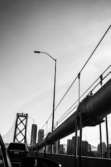 A black and white photograph of a suspension bridge with large cables and structures. Cars are visible on the roadway, and the skyline of a city with tall buildings can be seen in the background. The layout suggests an urban environment with a clear sky above.