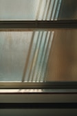 An abstract composition of light and shadows is visible through textured glass. The sunlight filters through, casting soft, elongated shadows, creating an interplay of patterns on the surface. Horizontal lines of metal frames divide the glass, contributing to a minimalist yet dynamic appearance.