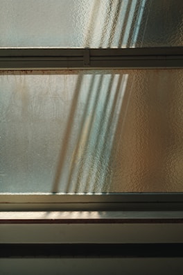 An abstract composition of light and shadows is visible through textured glass. The sunlight filters through, casting soft, elongated shadows, creating an interplay of patterns on the surface. Horizontal lines of metal frames divide the glass, contributing to a minimalist yet dynamic appearance.