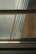 An abstract composition of light and shadows is visible through textured glass. The sunlight filters through, casting soft, elongated shadows, creating an interplay of patterns on the surface. Horizontal lines of metal frames divide the glass, contributing to a minimalist yet dynamic appearance.