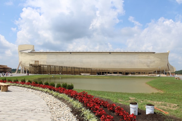 A large wooden ship, resembling a historical ark, is situated in the backdrop of a landscaped area. A pathway made of stone tiles leads towards the ark, with a line of vibrant red flowers and green shrubbery on one side. A small pond mirrors the ark, while the sky overhead is partly cloudy, adding to the serene setting.