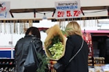 Two people are standing in front of a market stall. The person on the right is holding a large bouquet of flowers wrapped in brown paper. The stall features hanging necklaces and offers watch battery replacement services, as indicated by the sign above.