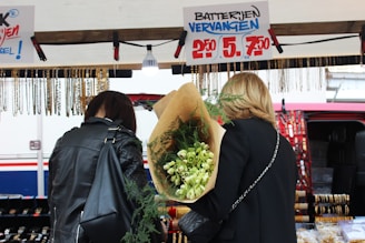 Two people are standing in front of a market stall. The person on the right is holding a large bouquet of flowers wrapped in brown paper. The stall features hanging necklaces and offers watch battery replacement services, as indicated by the sign above.