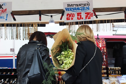 Two people are standing in front of a market stall. The person on the right is holding a large bouquet of flowers wrapped in brown paper. The stall features hanging necklaces and offers watch battery replacement services, as indicated by the sign above.