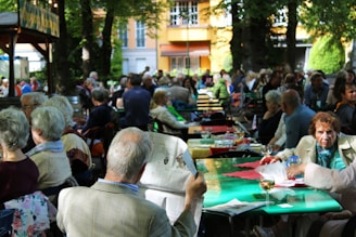 A busy outdoor beer garden filled with people sitting at tables under the shade of trees. Most of the patrons appear to be older adults, socializing and enjoying drinks. A few are seen reading newspapers, and there is a relaxed, convivial atmosphere. Colorful tablecloths cover the tables, and the area is adorned with lush green foliage.