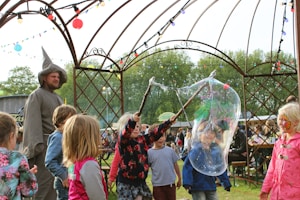 Children are playing with large soap bubbles at an outdoor festival. A person dressed as a wizard stands nearby, surrounded by several kids with face paint. Strings of colorful lights hang above, and a crowd of people can be seen in the background, sitting at tables amidst a green, leafy setting.