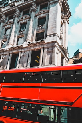 A vibrant red double-decker bus is passing by a classic, ornate building with large windows and detailed stonework. The architecture showcases tall columns and decorative elements. The sky is clear with patches of blue.