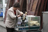 A person is preparing sugarcane juice using a manual juicer next to a stack of door panels. The individual is wearing gloves while feeding the sugarcane into the machine. A transparent container beside holds more sugarcane sticks. The setting appears to be an outdoor market or street setup.