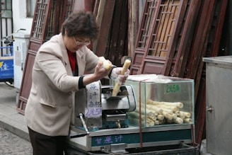 A person is preparing sugarcane juice using a manual juicer next to a stack of door panels. The individual is wearing gloves while feeding the sugarcane into the machine. A transparent container beside holds more sugarcane sticks. The setting appears to be an outdoor market or street setup.