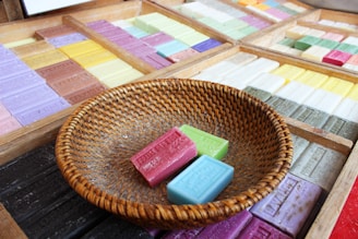 A variety of colorful soap bars are neatly arranged in wooden trays. In the foreground, a round woven basket contains three bars of soap in pink, green, and blue. Each bar displays embossed text, indicating different fragrances.