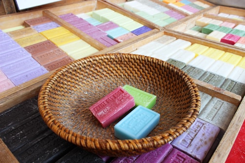 A variety of colorful soap bars are neatly arranged in wooden trays. In the foreground, a round woven basket contains three bars of soap in pink, green, and blue. Each bar displays embossed text, indicating different fragrances.