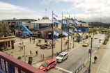 A bustling outdoor area featuring Pier 39 with several flags waving in the wind. People are walking around, and numerous shops and attractions are visible, including a blue slide. There are parked vehicles and taxis on the street adjacent to the pier. Trees and lampposts line the street, while the sky is partly cloudy.