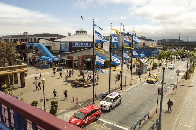 A bustling outdoor area featuring Pier 39 with several flags waving in the wind. People are walking around, and numerous shops and attractions are visible, including a blue slide. There are parked vehicles and taxis on the street adjacent to the pier. Trees and lampposts line the street, while the sky is partly cloudy.