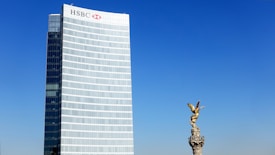A tall modern skyscraper with the HSBC logo stands prominently against a clear blue sky. Adjacent to it, a golden statue of an angel is perched atop a stone column. The architectural elements are distinct, with the smooth glass facade of the building contrasting with the detailed sculpture of the statue.