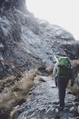 A person with a large green backpack and hiking gear ascends a rugged mountain path surrounded by rocky terrain and sparse vegetation.