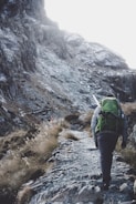 A person with a large green backpack and hiking gear ascends a rugged mountain path surrounded by rocky terrain and sparse vegetation.