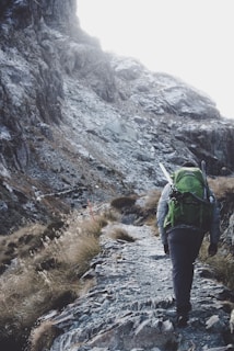 A person with a large green backpack and hiking gear ascends a rugged mountain path surrounded by rocky terrain and sparse vegetation.