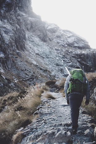 A person with a large green backpack and hiking gear ascends a rugged mountain path surrounded by rocky terrain and sparse vegetation.