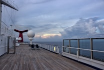A wooden deck of a cruise ship stretches out towards the horizon under a vast, cloudy sky. The deck is lined with glass barricades on one side, providing a clear view of the ocean. Loungers are placed sparsely along the deck. Prominently featured is a large red funnel with a wing-like structure and several white spherical structures, possibly serving as radar or communication equipment. The atmosphere appears calm and serene.