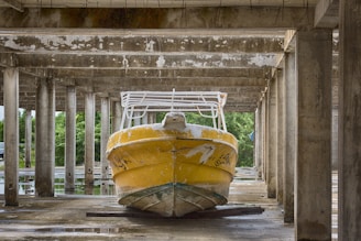 A yellow boat with visible wear and peeling paint is stored under a concrete structure with multiple columns. Surrounding the area are trees and greenery visible in the background.