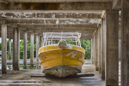 A yellow boat with visible wear and peeling paint is stored under a concrete structure with multiple columns. Surrounding the area are trees and greenery visible in the background.