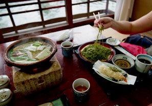 A cozy setting featuring a traditional Japanese meal with a bubbling hot pot filled with vegetables and tofu, accompanied by a plate of green soba noodles being lifted with chopsticks. Assorted tempura and dipping sauces complete the meal, set against a wooden table with an intricate design on the hot pot base.