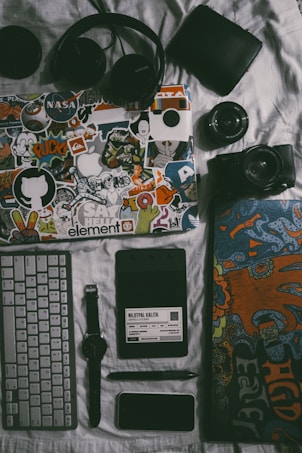 A collection of personal items including a laptop covered in colorful stickers, a pair of black headphones, a black wallet, a camera with a lens, a keyboard, a wristwatch, a notepad with a pen, and a smartphone. The items are arranged on a white surface, giving a sense of organization and personal style.
