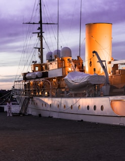 A large, elegant ship docked at a harbor during sunset, with its exterior lights casting a warm glow. The ship features multiple decks, round portholes, and a large smokestack. A sailor in a white uniform stands on the dock nearby.